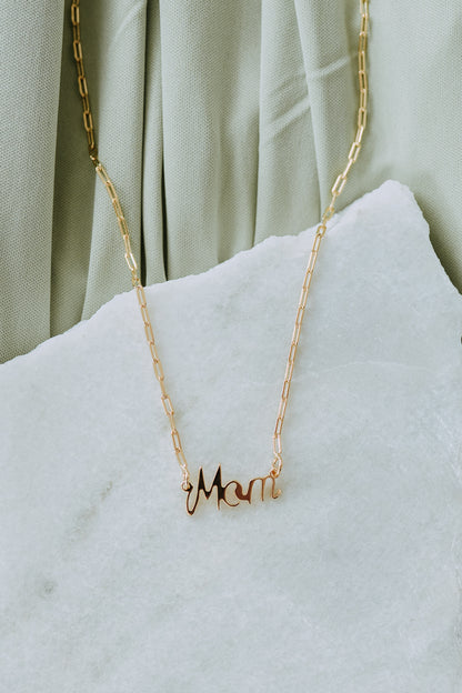 18k gold filled Mom necklace displayed on a white marble jewelry prop with soft green fabric.