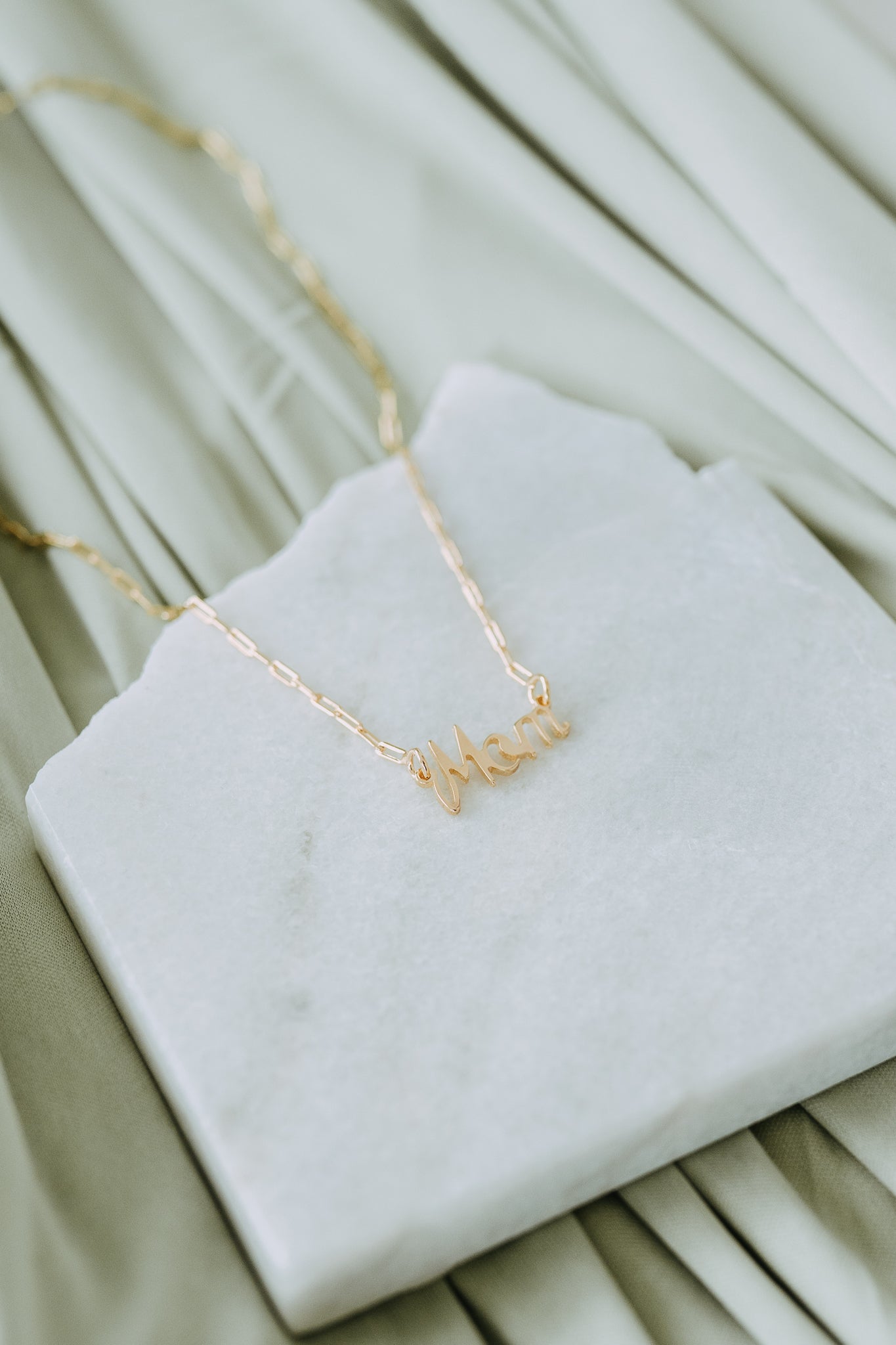 Close-up of a woman holding a gold Mom necklace featuring a delicate script pendant and paperclip chain.