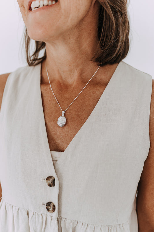 Sterling silver oval engraved locket necklace on a delicate box chain worn with a neutral linen top.