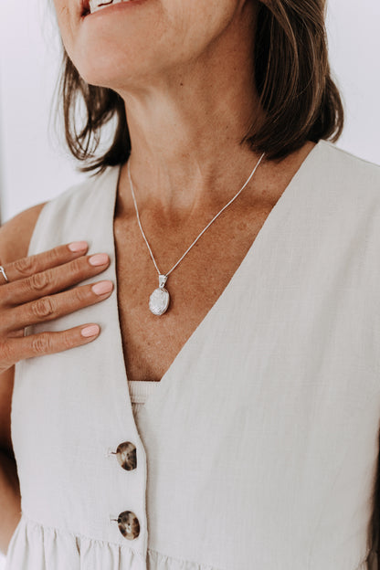 Sterling silver oval engraved locket necklace on a delicate box chain worn with a neutral linen top.