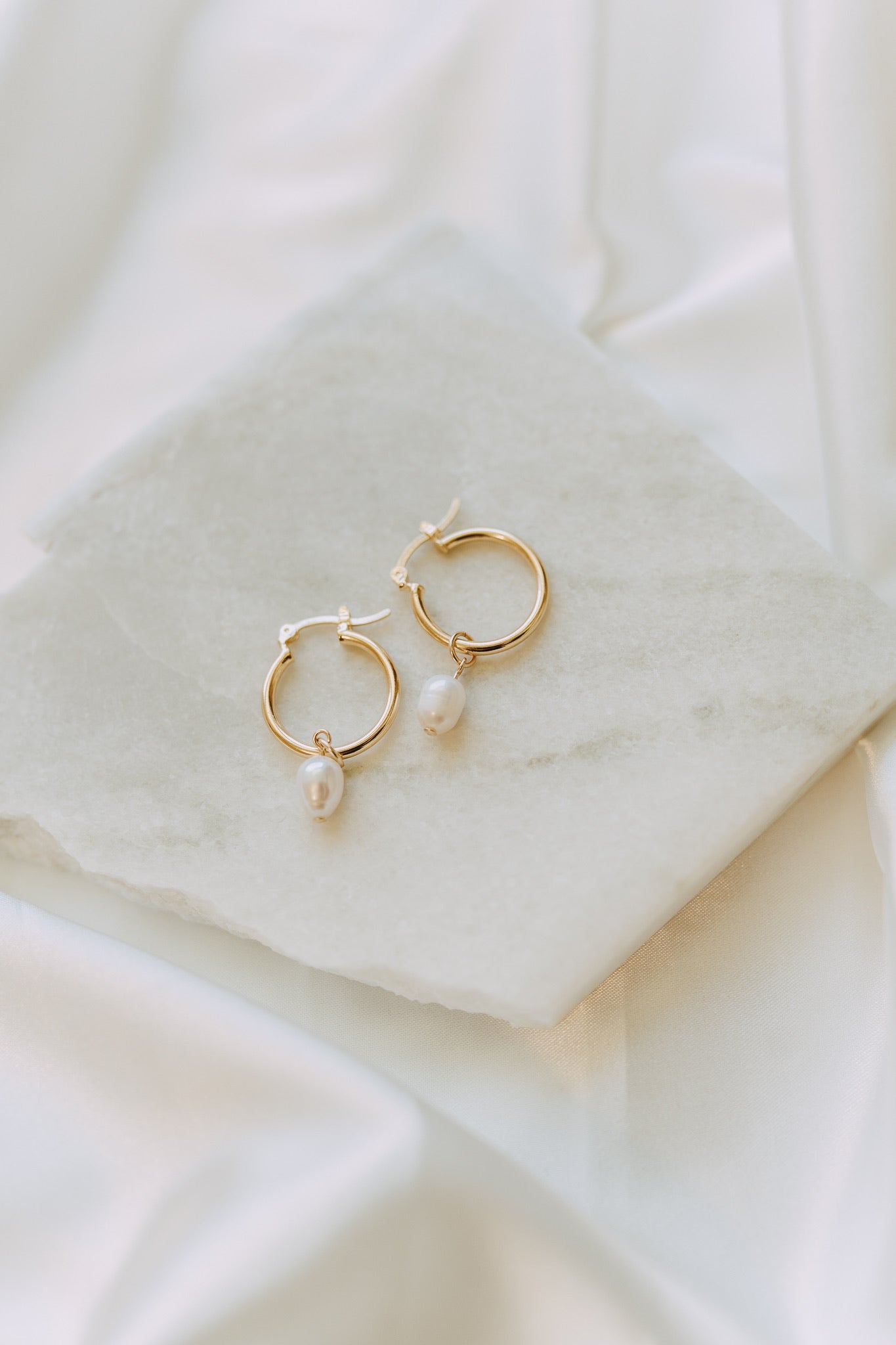 Minimal gold pearl drop hoop earrings displayed on white marble surface with soft neutral styling