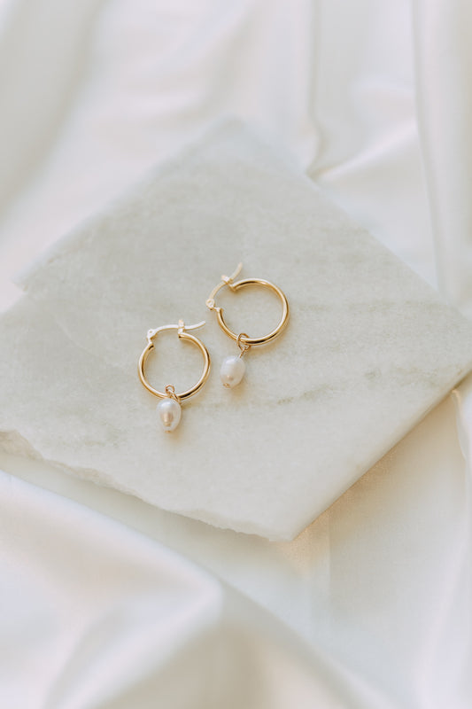 Minimal gold pearl drop hoop earrings displayed on white marble surface with soft neutral styling