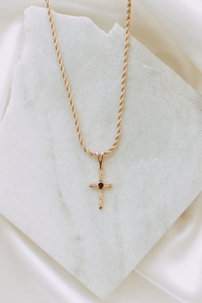 Flat lay of engraved gold cross pendant necklace on a twisted rope chain over white marble backdrop